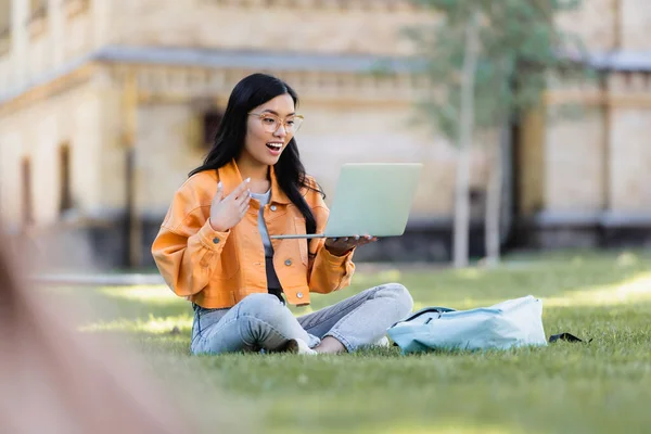 Happy asian woman gesturing during video chat on lawn in park — Stock Photo