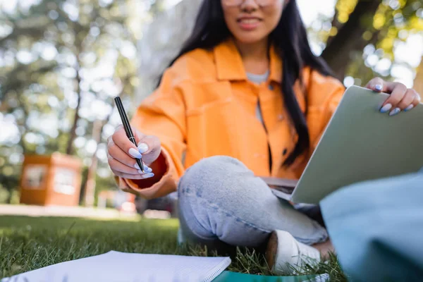 Cropped view of blurred student sitting on lawn with pen and laptop — Stock Photo
