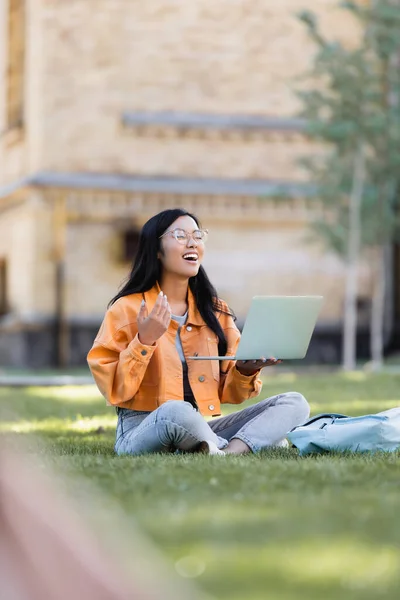 Cheerful asian student laughing with closed eyes during video chat on lawn in park — Stock Photo