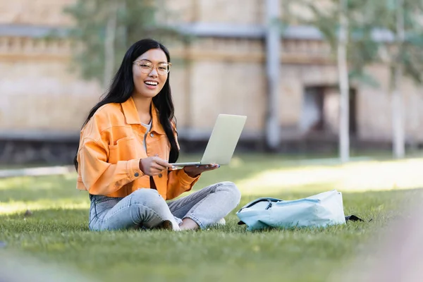 Glückliche Asiatin sitzt mit Laptop auf Gras und lächelt in die Kamera — Stockfoto