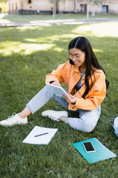 Feliz asiático mujer usando digital tablet cerca smartphone y notebooks en hierba - foto de stock