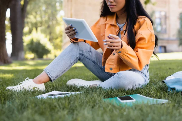 Vista cortada do estudante em jaqueta laranja usando tablet digital na grama no parque — Fotografia de Stock