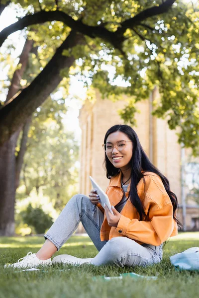 Happy asian student in eyeglasses looking at camera while sitting on lawn with digital tablet — Stock Photo