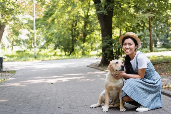 Cheerful asian woman in straw hat looking at camera while hugging labrador in park — Stock Photo