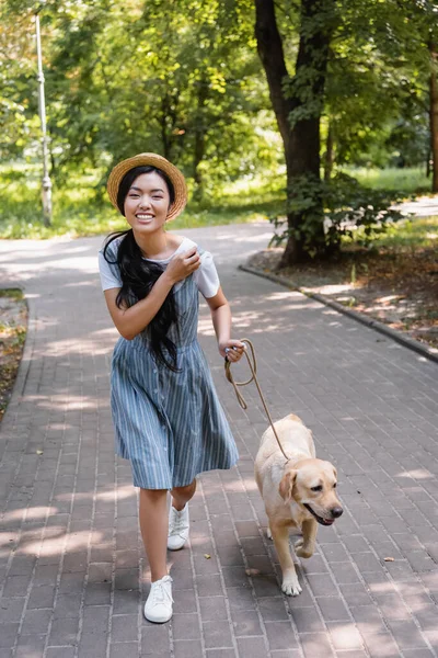 Excited asian woman smiling at camera while strolling with dog in park — Stock Photo