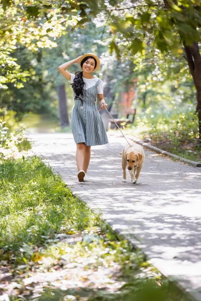 Cheerful asian woman adjusting straw hat while walking with yellow labrador — Stock Photo