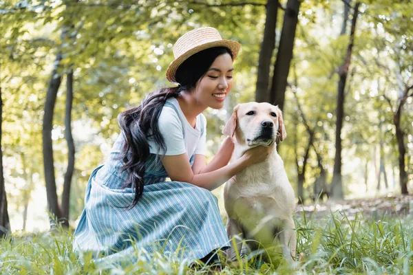 Giovane donna asiatica in cappello di paglia coccole giallo labrador nel parco — Foto stock