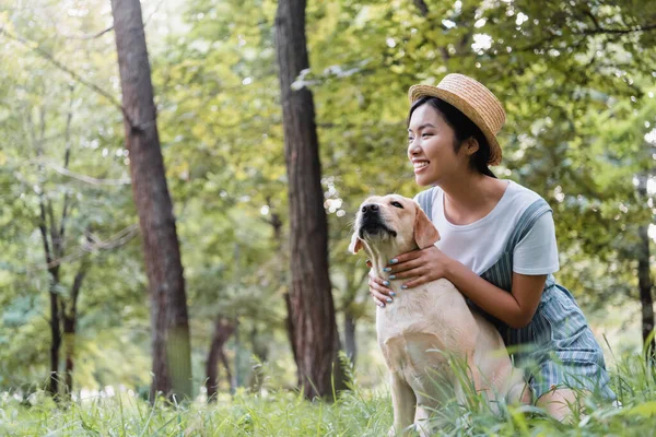 Heureux asiatique femme en paille chapeau étreignant chien et regardant loin dans parc — Photo de stock