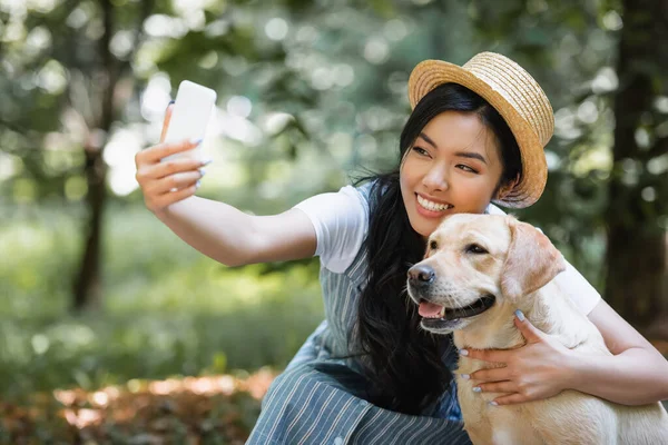 Alegre asiático mujer en paja sombrero tomando selfie con amarillo labrador en parque - foto de stock