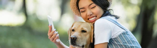 Веселая азиатка с закрытыми глазами, делающая селфи с собакой на открытом воздухе, баннер — стоковое фото