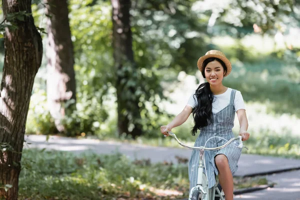 Веселая азиатка в соломенной шляпе, улыбающаяся в камеру во время езды на велосипеде в парке — стоковое фото
