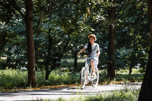 Joven asiático mujer en paja sombrero y sundress ciclismo en parque - foto de stock
