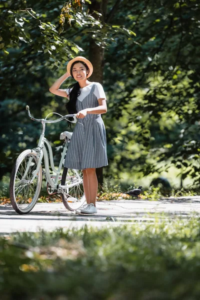 Asiático mulher tocando palha chapéu enquanto olhando para longe perto de bicicleta no parque — Fotografia de Stock