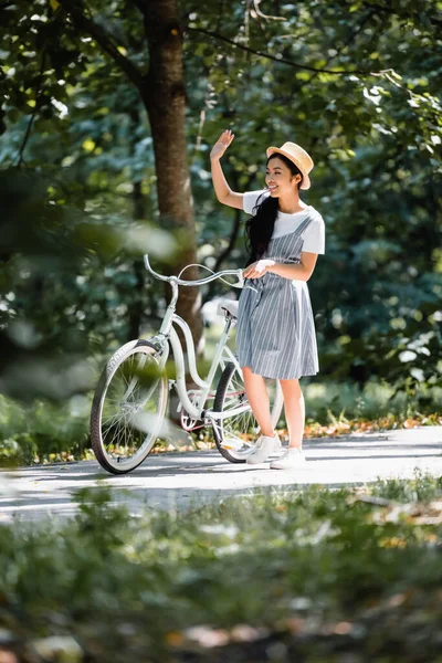 Complacido asiático mujer mirando lejos y agitando mano cerca de bicicleta en parque - foto de stock