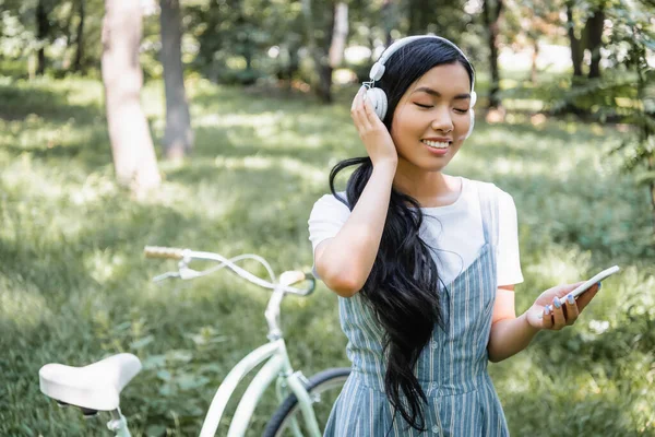 Усміхнена азіатська жінка регулює навушники під час використання мобільного телефону біля розмитого велосипеда — стокове фото