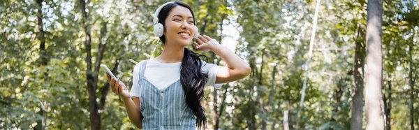 Mujer asiática feliz con teléfono inteligente mirando hacia otro lado mientras escucha música en el parque, pancarta - foto de stock