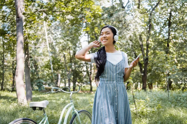 Joyful asian woman with mobile phone listening music in headphones near bicycle — Stock Photo