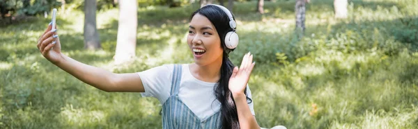 Heureux asiatique femme dans casque agitant la main tout en prenant selfie dans parc, bannière — Photo de stock