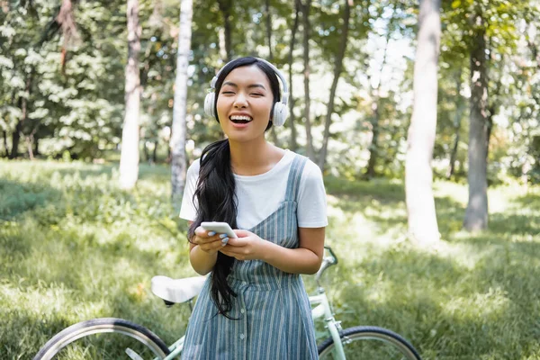 Excitado mulher asiática com smartphone rindo em fones de ouvido perto de bicicleta turva — Fotografia de Stock