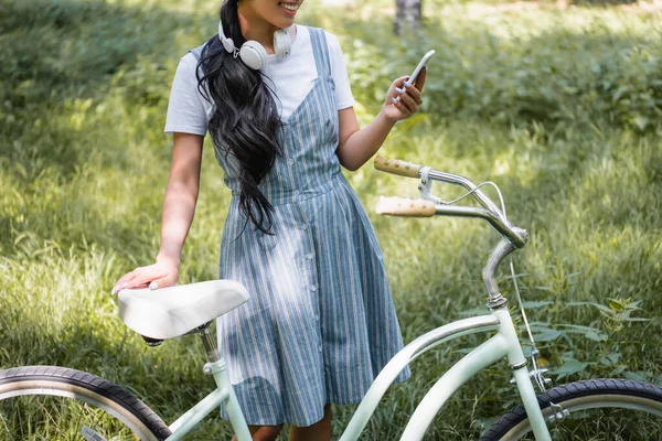 Vue recadrée de la femme souriante avec des messages écouteurs sur téléphone portable près de vélo — Photo de stock