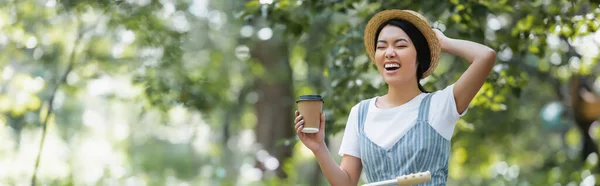 Excited asian woman with takeaway drink laughing with closed eyes in park, banner — Stock Photo