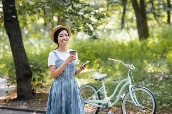 Счастливая азиатская женщина с напитком на вынос и мобильный телефон глядя на камеру возле велосипеда в парке — стоковое фото