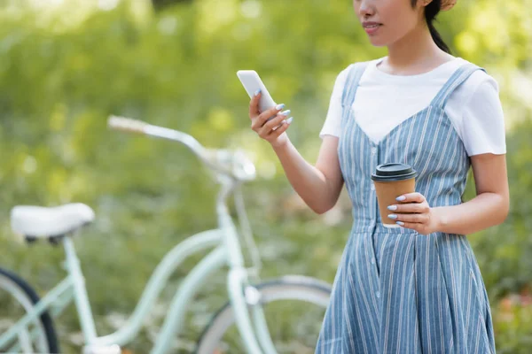 Cropped view of woman with takeaway drink chatting on smartphone near blurred bicycle — Stock Photo