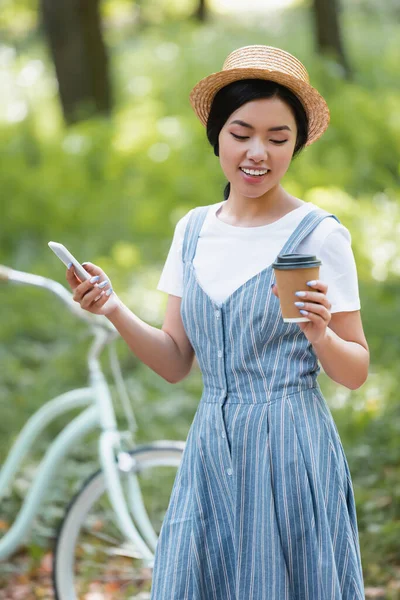 Mujer asiática complacida con teléfono inteligente y café para ir de pie cerca de la bicicleta en el parque - foto de stock