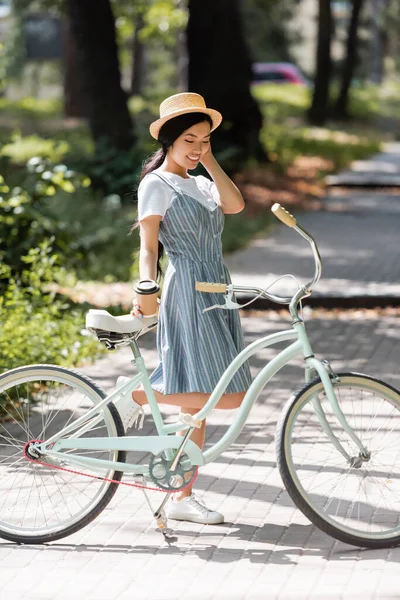 Satisfeito ásia mulher no listrado sundress pé perto de bicicleta no parque — Fotografia de Stock