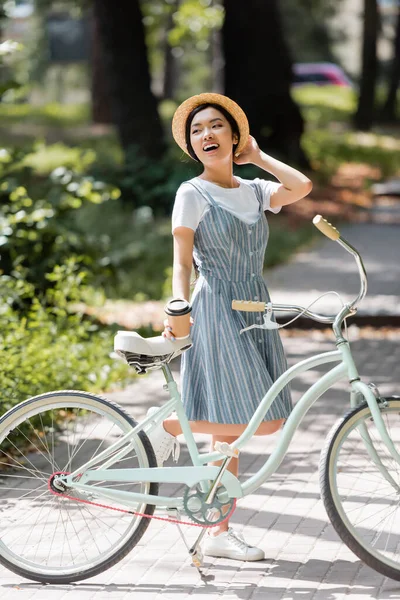 Rindo asiático mulher tocando palha chapéu enquanto olhando afastado perto de bicicleta no parque — Fotografia de Stock