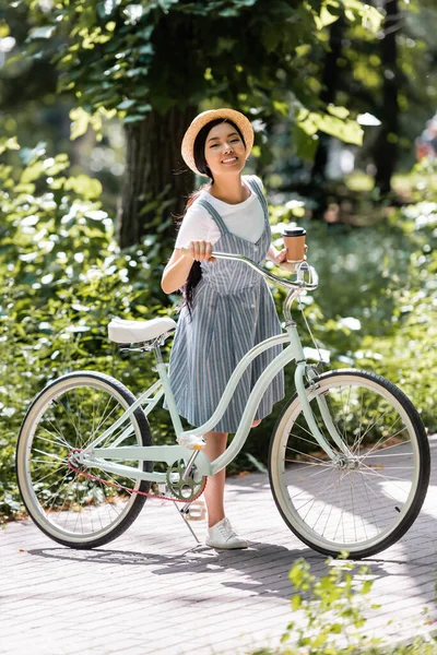 Pleine longueur vue de heureux asiatique femme avec à emporter boisson et vélo dans le parc — Photo de stock