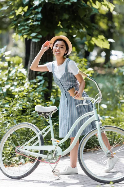 Young asian woman adjusting straw hat while smiling at camera near bike in park — Stock Photo