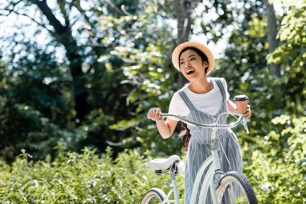 Happy asian woman with paper cup looking away and laughing near bike in park — Stock Photo