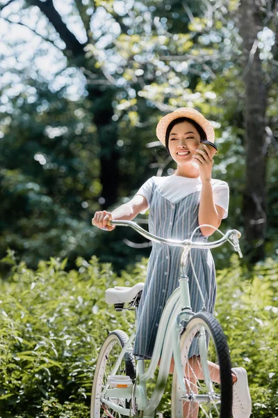 Счастливая азиатская женщина держит бумажную чашку возле лица, стоя с велосипедом в парке — стоковое фото