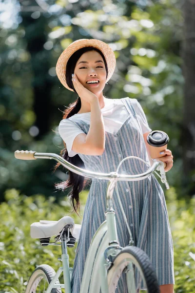 Alegre asiático mulher com café para ir sorrindo para câmera perto de bicicleta no parque — Fotografia de Stock