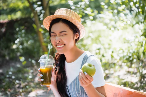 Joyful asian woman with closed eyes sitting in park with fresh apple and juice — Stock Photo