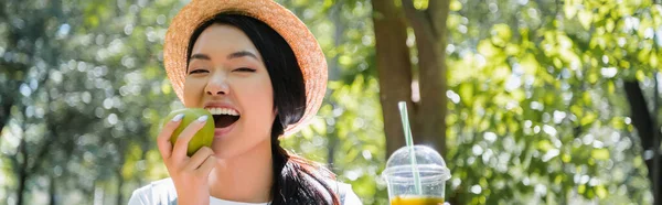 Positivo asiatico donna holding fresco succo mentre mangiare maturo mela in parco banner — Foto stock