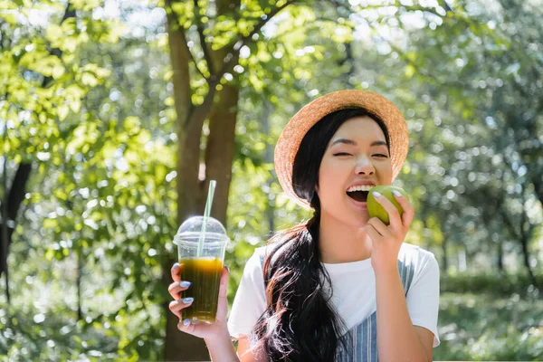 Heureux asiatique femme avec frais smoothie mordre juteux pomme dans parc — Photo de stock