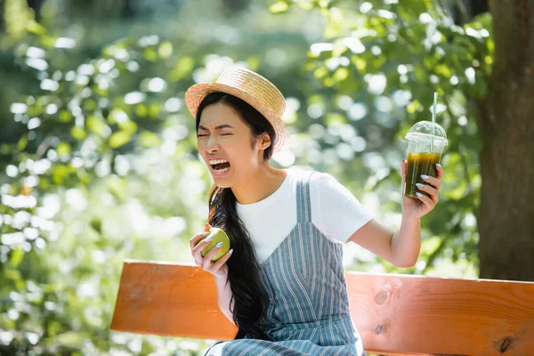 Asiático mujer con maduro manzana sensación disgustado mientras celebración de plástico taza de smoothie - foto de stock