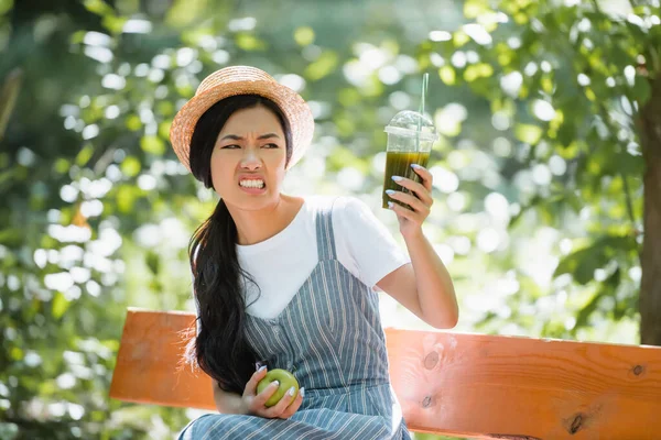 Disgustado asiático mujer mueca mientras mirando smoothie en plástico taza - foto de stock