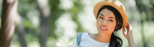 Young asian woman adjusting straw hat and smiling outdoors, banner — Stock Photo