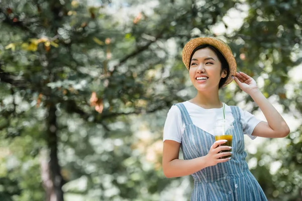 Cheerful asian woman with plastic cup of smoothie adjusting straw hat while looking away in park — Stock Photo