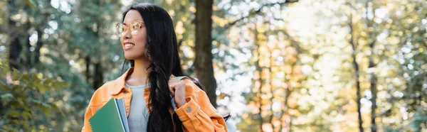 Asiatico studente in occhiali tenendo notebook e guardando lontano in parco, striscione — Foto stock