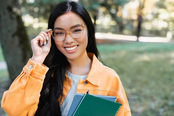 Brunette asian student adjusting eyeglasses while holding notebooks in park — Stock Photo
