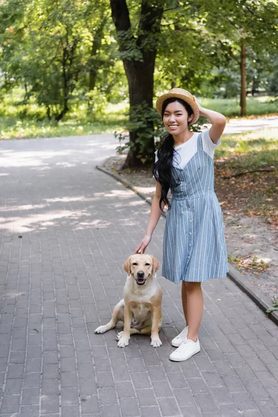 Joyeuse femme asiatique en robe de soleil et chapeau de paille regardant la caméra près de labrador dans le parc — Photo de stock