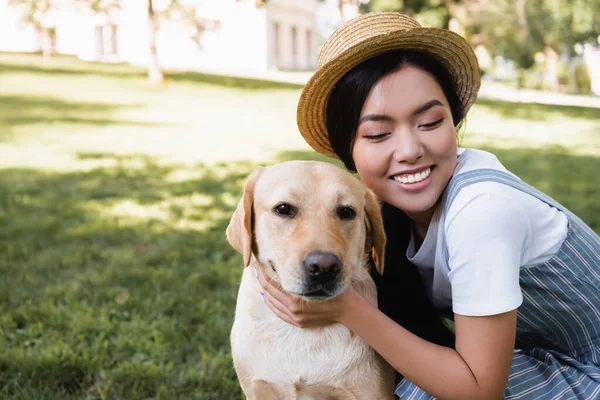 Heureux asiatique femme en paille chapeau embrassant jaune labrador dans parc — Photo de stock