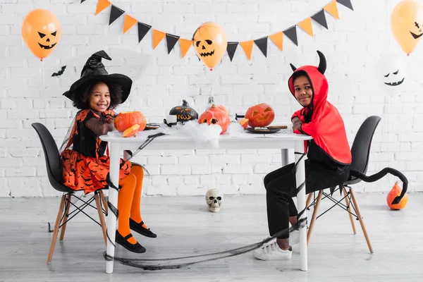 Щасливі афроамериканські діти в костюмах Хеллоуїна, сидячи за столом з гарбузами та прикрасами — стокове фото