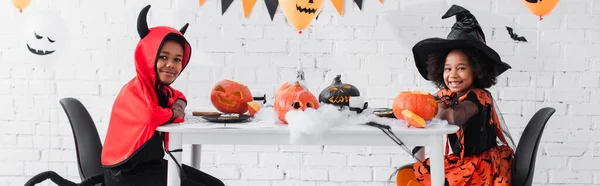 Crianças americanas africanas felizes em trajes de Halloween sentados à mesa com abóboras e decoração, banner — Fotografia de Stock