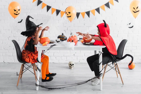 Spooky african american kids in halloween costumes sitting at table with pumpkins and decoration during party — Stock Photo