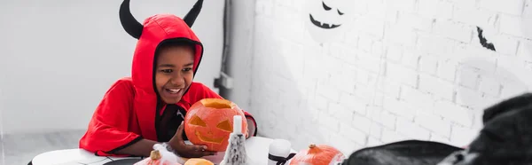 Happy african american boy in halloween costume holding carved pumpkin at home, banner — Stock Photo